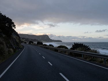 A wide, moody environmental photograph of a quiet New Zealand coastal highway at dawn, soft overcast daylight, deep navy and slate tones, 35mm