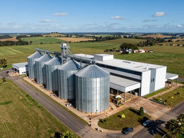 Aerial wide-angle view of a modern Brazilian farm headquarters with massive silver grain silos, paved transport roads, and green fields under a clear blue sky