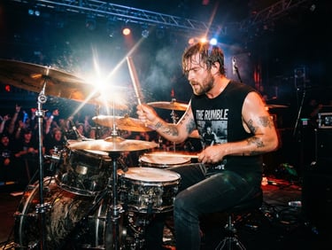 Dynamic action shot of drummer Matt Del Grosso mid-performance, drumsticks blurred in motion, cymbal flare under intense backlighting, gritty concert texture.