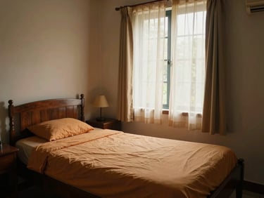 Warm Kerala care home room interior, afternoon light through a curtained window onto a neatly made single bed with ochre linen, wooden bedside table with a small lamp, modest and dignified, no residents present, eye-level wide shot