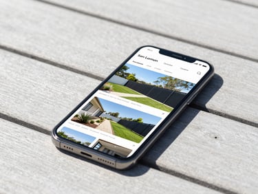 A smartphone showing a modern landscaping and fencing gallery website, resting on a clean timber deck, bright outdoor daylight, sharp focus