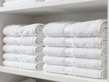 Brightly lit modern hair salon, neatly folded white towels stacked on a clean shelf, crisp daylight, sharp focus on the textiles.