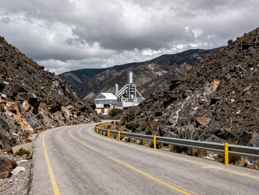 Wide environmental shot of an industrial road winding through a rugged mountain pass in Zacatecas, Mexico, with a silver mining facility visible in the distance under a dramatic overcast sky.