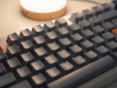 A close-up of a custom mechanical keyboard with matte grey keycaps, one warm orange accent key, soft moody desk lamp lighting.