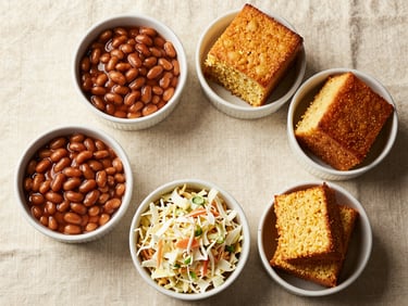 Overhead flat-lay of classic BBQ side dishes in white ceramic bowls on a cream linen surface — baked beans, coleslaw, cornbread — warm natural window light