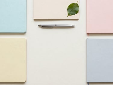 Top-down flat lay of neatly organized pastel notebooks, a sleek pen, and a single green leaf on a light cream desk, soft natural daylight, desaturated tones, 35mm