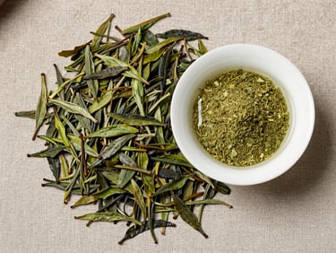 Flat-lay of loose green tea leaves and a small dish of EGCG extract powder on warm gray linen, diffused natural daylight from above, tight overhead crop, no bottles or labels