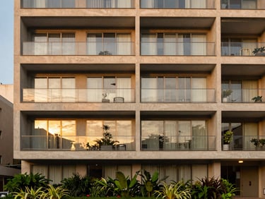 Warm architectural photography of a modern apartment building facade in São Paulo, soft golden-hour light reflecting on large glass windows, green landscaping below, 35mm