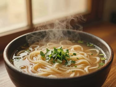 Warm editorial food photography of steaming noodle soup in a dark clay bowl, green herbs, soft golden-hour window light