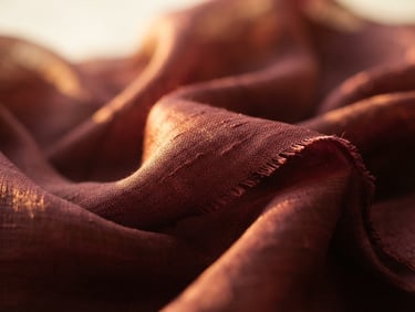 Macro photograph of deep maroon silk fabric draped elegantly, catching soft golden directional light, showing rich texture and weave