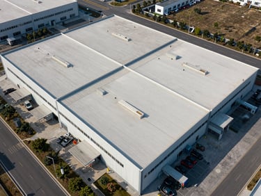 Diagonal approach aerial of a commercial campus or industrial facility, structured geometry of rooftops and parking visible, clear midday daylight, environmental context showing road access and surrounding land use