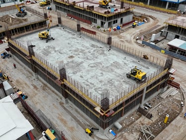 Overhead diagonal aerial of an active commercial construction site, concrete foundation and steel framing visible, equipment and workers small in scale to show site context, natural overcast daylight for accurate documentation
