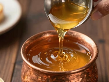Close-up of golden desi ghee in a copper vessel being tilted to pour a slow amber stream, hands visible at the rim, warm daylight from the left, dark wooden surface beneath
