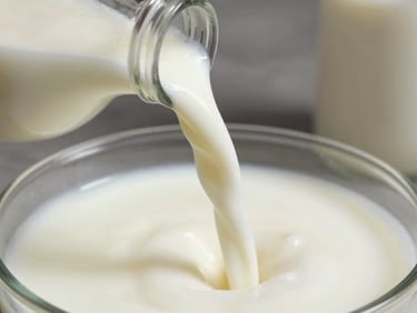 Intimate close-up of fresh whole milk being poured from a steel ladle into a clear glass bottle, natural daylight catching the white stream mid-pour, cream-coloured surface beneath, no background clutter