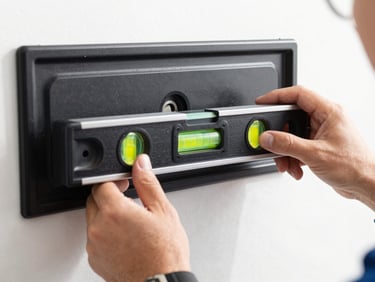 Close-up of a technician's hands using a bubble level on a sleek black metal TV wall bracket, bright natural daylight, sharp focus on the level indicator