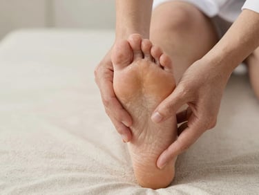 Close-up of therapist thumbs pressing into the sole of a foot with deliberate precision, soft diffused studio light, neutral warm tones, clean linen background, clinical-calm framing