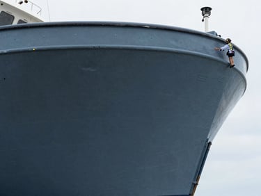 Detailed shot of a damaged boat hull, surveyor pointing to a specific area, natural overcast daylight, cool slate and deep blue tones, sharp focus, 35mm