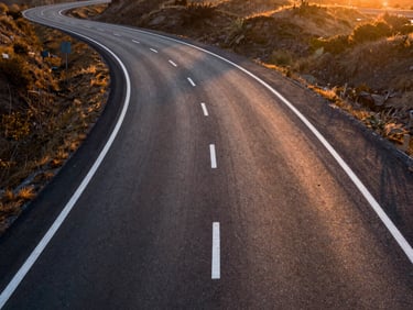 Aerial high-angle shot of a newly paved asphalt highway curving smoothly through hills, sharp white lane markings, sunset light