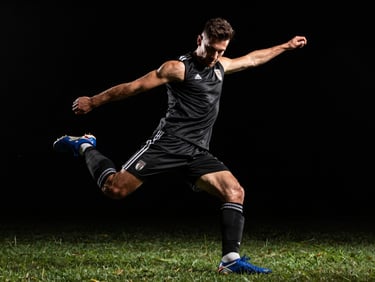 Low-angle wide shot of a soccer player mid-strike, dramatic side-lighting highlighting muscle definition, pitch-black background.