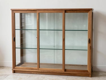 Solid teak glass-door display cabinet against a white wall under even daylight, glass panels clear, teak frame grain visible, interior shelves empty, pale stone floor