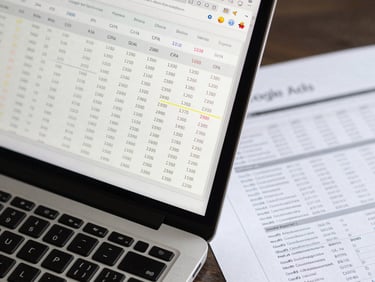 Extreme close-up overhead flat-lay of a laptop screen displaying a Google Ads dashboard with highlighted CPA column, cool white studio light, sharp focus on the numeric data rows, printed report with annotated KPIs beside the keyboard