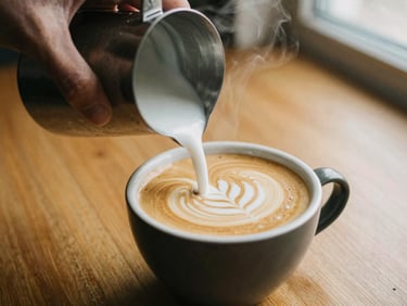A close-up of a hand pouring steamed milk into a ceramic cup of espresso, creating latte art, warm timber background