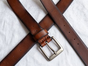 Overhead flat-lay of a wide leather belt with a brushed silver buckle on white linen, bright fill light showing grain texture and punched holes detail