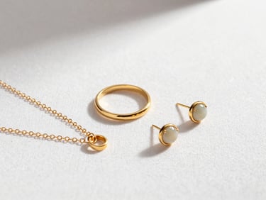 Close-up flat-lay of fine jewelry pieces arranged on white linen — a delicate gold chain, a minimal ring, a small stud earring — bright controlled studio light highlighting metal surface and stone setting clarity