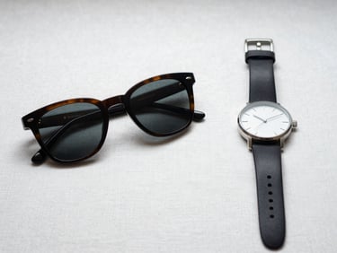 Flat-lay of sunglasses and a minimal watch placed side by side on white linen, controlled studio light catching lens quality and case finish, tight framing revealing hinge and strap material detail