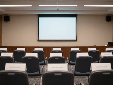 Wide shot of a conference room setup from the back, rows of empty chairs with delegate nameplates, projector screen at the front, clean institutional lighting