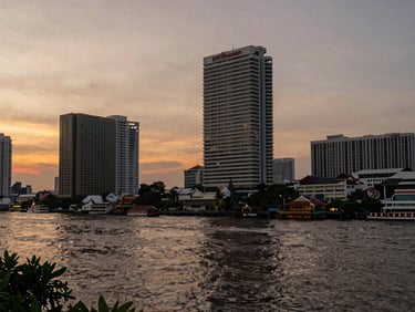 Riverside Bangkok at dusk, view from a Sathorn-adjacent vantage looking toward the Chao Phraya, warm ambient light, water and towers in frame, no people