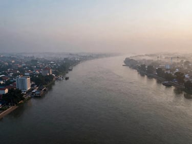 Aerial view looking north along the Chao Phraya river at dawn, low mist over water, riverside towers emerging from haze, wide environmental frame, no people