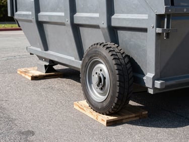 Close-up of a heavy-duty steel dumpster wheel resting on thick protective wooden boards on a clean asphalt driveway, sharp industrial photography, high contrast