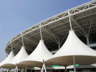 Low-angle architectural photography of a massive white tensile membrane structure over a modern stadium, bright daylight, steel trusses visible, clear sky