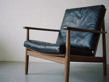 Architectural furniture photography, low-angle shot of a minimalist lounge chair in American walnut and full-grain leather, soft side daylight, deep navy shadows.