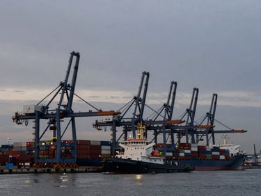 Wide landscape view of a busy commercial harbor at dusk, industrial cranes silhouetted against a steel-gray sky, a heavy tugboat guiding a container ship.
