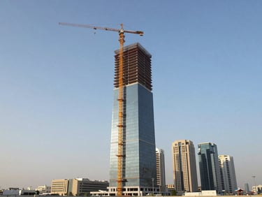 Wide environmental shot of a high-rise commercial tower under construction in an urban Gulf city, tower crane at peak height, steel framework rising above surrounding buildings, clear blue sky, midday natural light