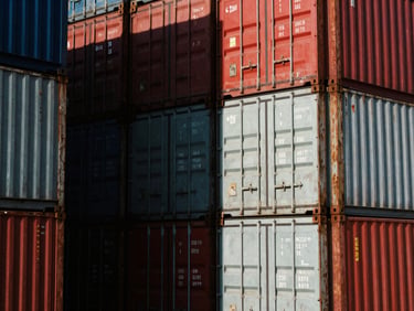 High-contrast close-up of heavy industrial shipping containers stacked at a deepwater port, dramatic shadows, crimson steel accents, 35mm film grain