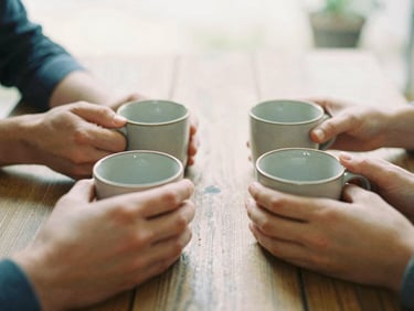 Close-up of hands holding warm ceramic mugs around a rustic wooden table, soft overcast daylight, natural and unposed, 35mm film style.