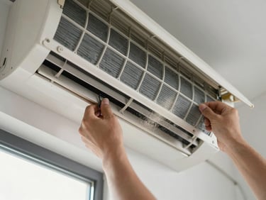 Close-up overhead shot of a technician's hands removing a dusty AC filter from a wall-mounted indoor unit, the filter's aluminum mesh grid clearly visible, daylight from a nearby window illuminating the fine dust particles mid-air, clinical and detailed