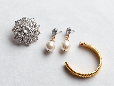 Overhead flat-lay of fine jewelry — a diamond brooch, pearl drop earrings, and a gold Art Deco bracelet — arranged on white velvet under bright studio light