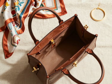 Overhead flat-lay of an open designer handbag showing interior leather stitching and hardware, beside folded vintage silk scarves and a gold bracelet on cream linen, bright north-facing daylight