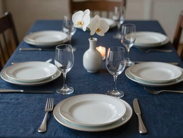 Close-up of an intimate private dining table set for eight — deep navy linen, hand-painted porcelain chargers, single white orchid stem in a slender vase, candlelight warmth, no guests, pre-service composition