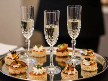 Close-up of a cocktail reception tray—three crystal champagne flutes partially filled, gold-rimmed canapés arranged precisely on a mirrored tray, soft warm ambient light