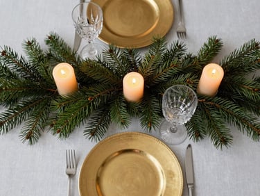 Overhead of a holiday table arrangement—deep green spruce boughs framing gold-rimmed plates, pillar candles casting warm glow, crystal glassware, white linen, no people