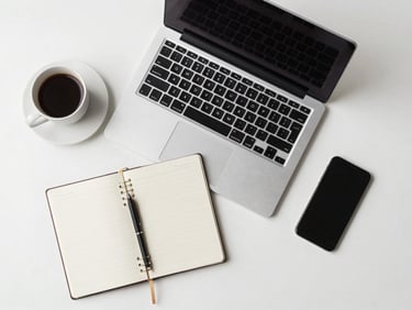 Overhead flat-lay of a modern desk workspace — open laptop, notebook, coffee cup, and phone arranged neatly on a white surface under diffused studio light, no people, clean and minimal