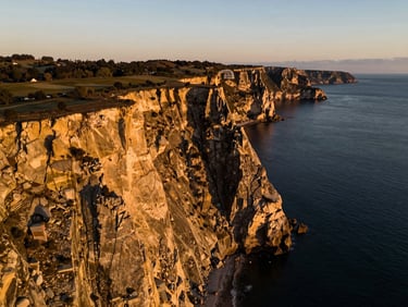 High-altitude drone photography of a premium coastal cliffside land plot, dramatic low-sun angle casting long shadows, rich gold and dark tones, wide-angle lens.