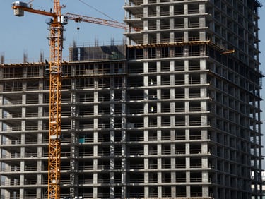 Wide shot of a high-rise building under construction, cranes in the background, workers in safety gear on scaffolding, sharp industrial photography, high-contrast natural daylight, deep navy and metallic tones, crisp focus, 35mm architectural lens.