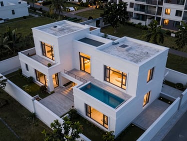 Drone overhead view of a completed luxury residential villa compound at dusk, clean white rendered facades with flat roof terraces, geometric pool visible, warm exterior lighting, no people