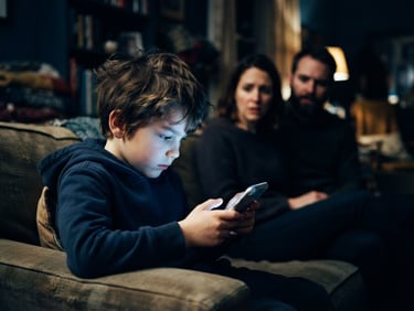 Candid emotional editorial photo of a young child staring intently at a glowing smartphone screen in a dimly lit room, worried parents in the soft-focus background looking on with concern, deep blue tones, high contrast.